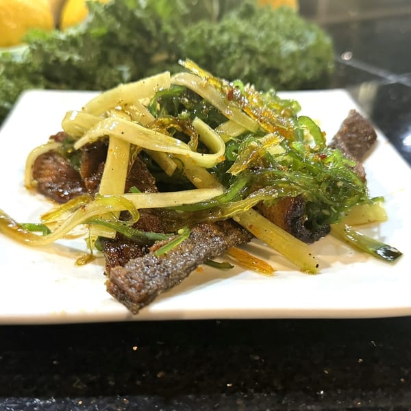 A white plate holds a sushi dish with dark meat, green seaweed, and yellow noodles.
