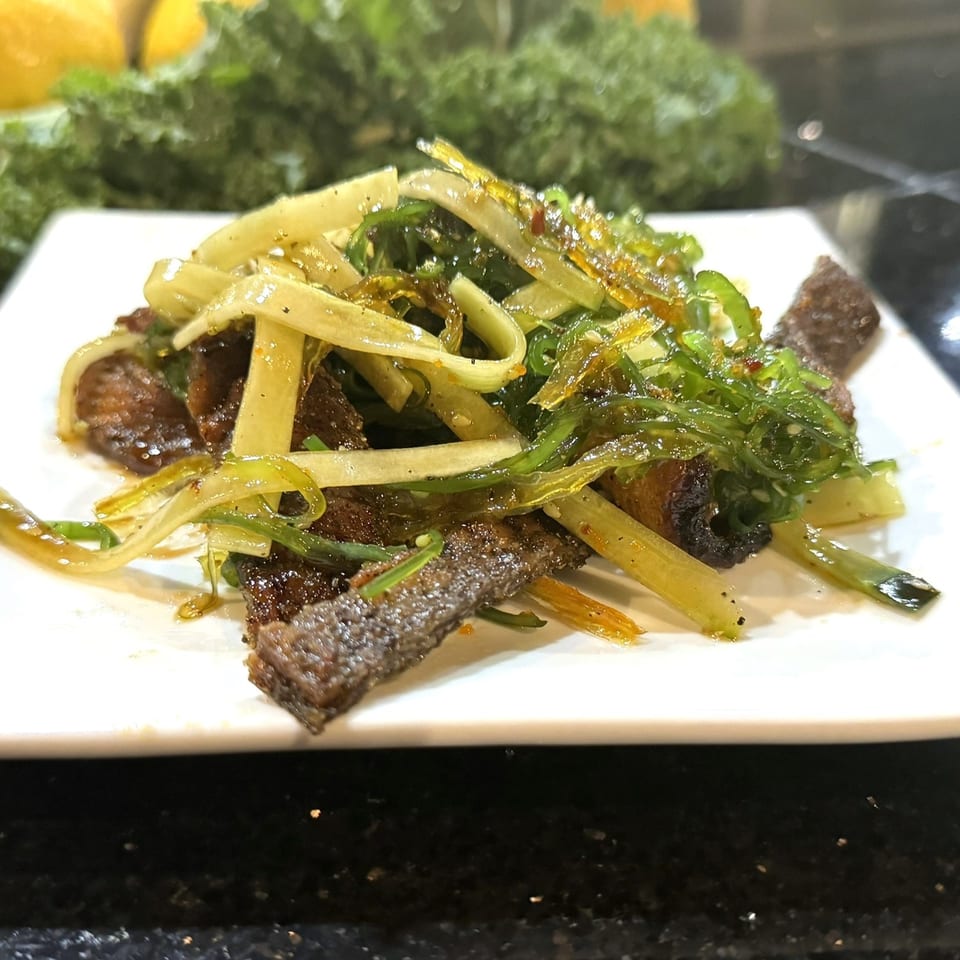 A white plate holds a sushi dish with dark meat, green seaweed, and yellow noodles.